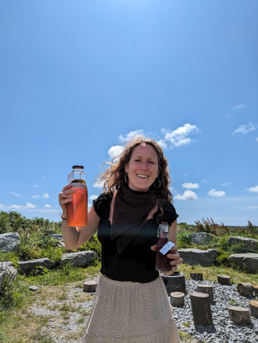Jen Doran standing in a stone circle on a sunny day smiling holding up bottles of herbal drints