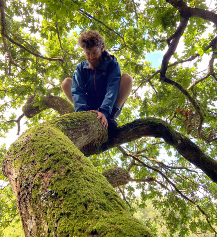 profile image of Luc O'Rourke looking thoughtful on a mossy tree trunk surrounded by leaves